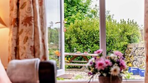 Habitació amb vista a un jardí a través d'una porta de vidre, una taula de fusta amb un ram de flors i un sofà.
