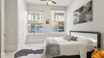 Quarto com cama, ventoinha no teto, duas janelas e tapete.
