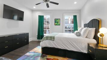 Un dormitorio con cama, ventilador de techo, televisor, mesita de noche con lámpara y ventana con cortinas.