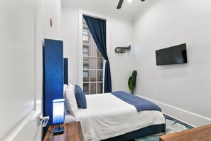 Un dormitorio con cama, lámpara de noche, televisión y ventana con cortinas.