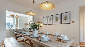 Ein Essbereich mit einem langen Holztisch, gedeckt für vier Personen, umgeben von gewobenen Pendelleuchten, gerahmten botanischen Drucken und Blick auf eine verschneite Landschaft.
