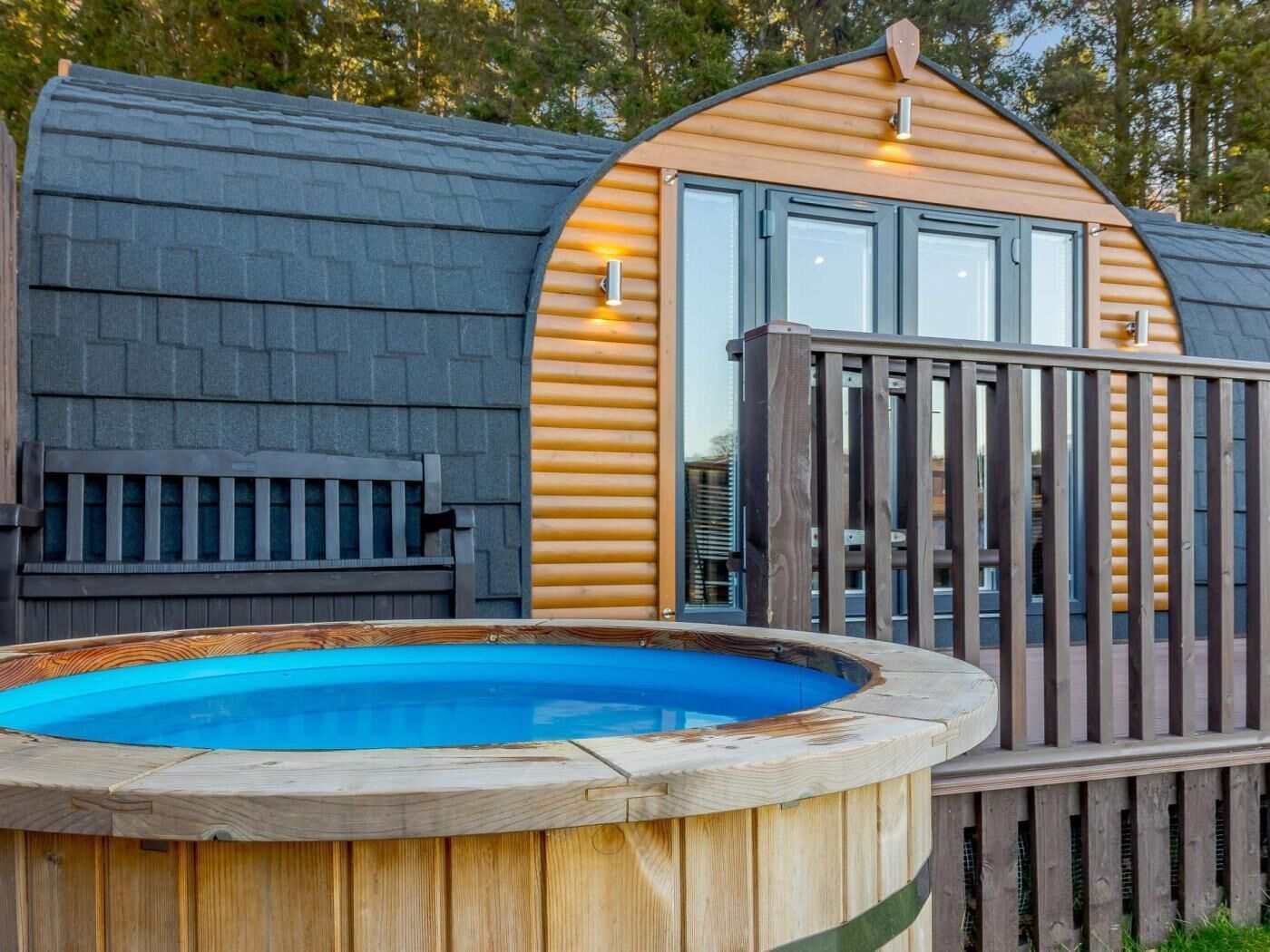 A wooden hot tub with a wooden deck and railing, a bench, and a building with a sloped roof and large windows.