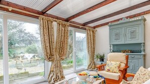 Een kamer met een houten plafond, een groot raam met gordijnen, een houten stoel, een tafel met een schaal fruit, en een kast.