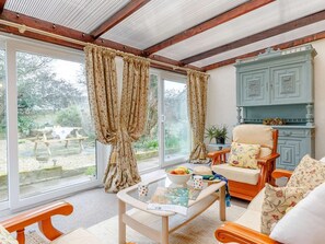 Ein Zimmer mit Holzboden, einem großen Fenster mit Vorhängen, einem Holzstuhl, einem Tisch mit Obstschale und einem Schrank.
