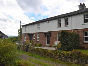 A two-story house with a stone wall and a brick extension, a front door, and a small porch with potted plants. - 2 Bed in Keswick (oc-sz250) (Braithwaite)
