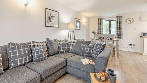 A modern living room with a grey sectional sofa, a dining area with a table and chairs, a lamp, and a clock on the wall. - 2 Bed in Woolacombe (oc-westd) (Woolacombe)