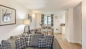 A living room with a sofa, a lamp, a dining area with a table and chairs, and a kitchen area with a microwave. - 2 Bed in Woolacombe (oc-westd) (Woolacombe)