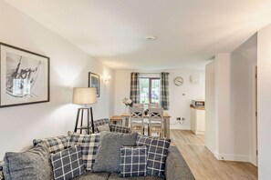 A living room with a sofa, a lamp, a dining area with a table and chairs, and a kitchen area with a microwave. - 2 Bed in Woolacombe (oc-westd) (Woolacombe)
