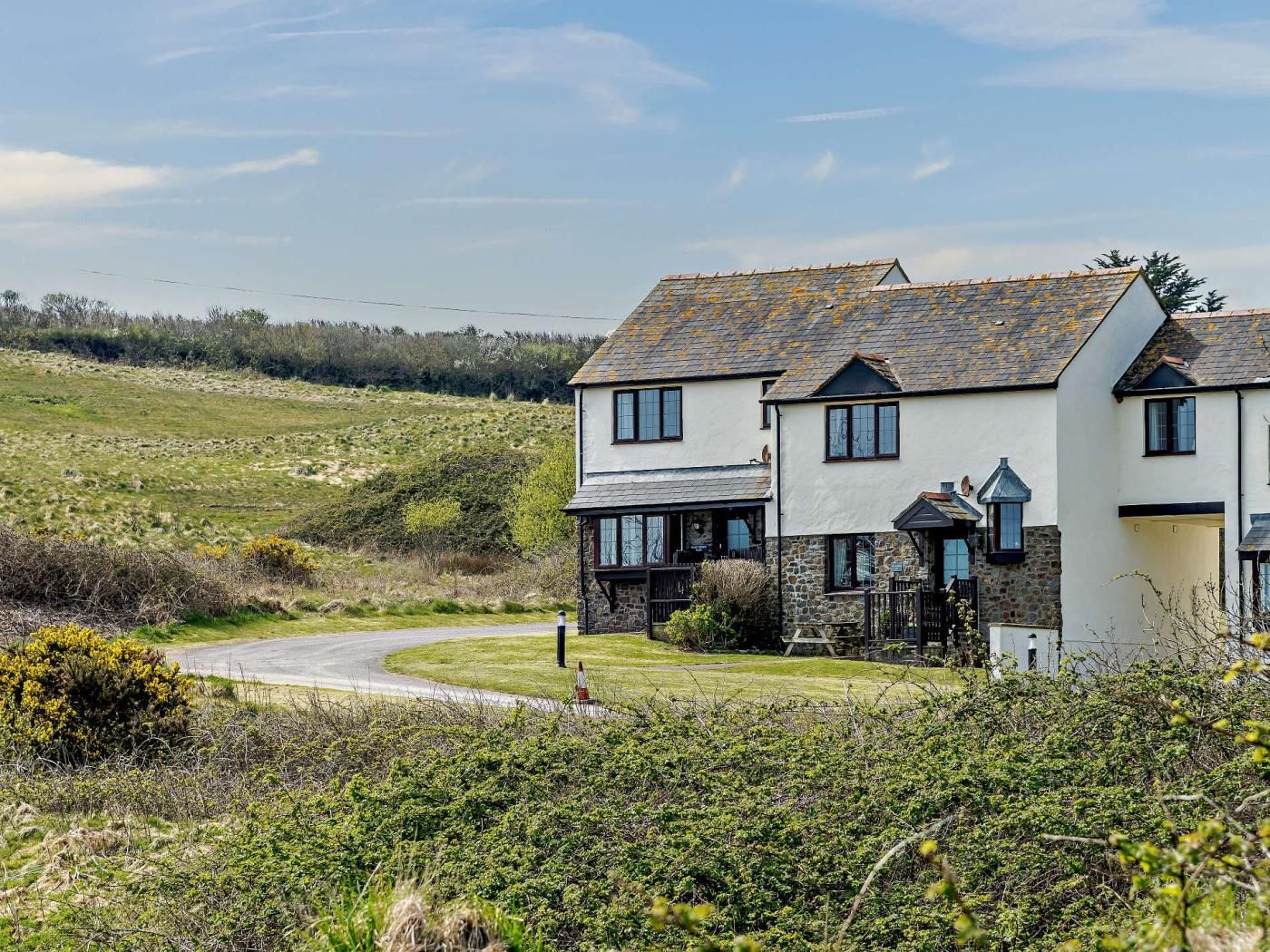 A two-story house with a stone foundation and a tiled roof, surrounded by a grassy landscape and a winding road.