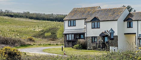 A two-story house with a stone foundation and a tiled roof, surrounded by a grassy landscape and a winding road.