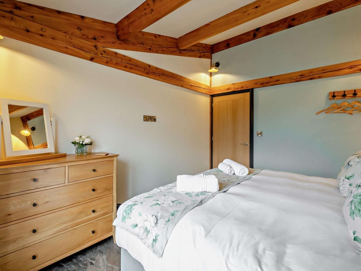 A bedroom with a wooden ceiling, a bed with floral bedding, a dresser, a mirror, and a door.