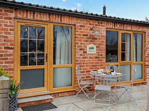 Ein Backsteinhaus mit großen, holzrahmenverzierten Fenstern und einer kleinen Terrasse mit Metallstühlen und einem Tisch.
