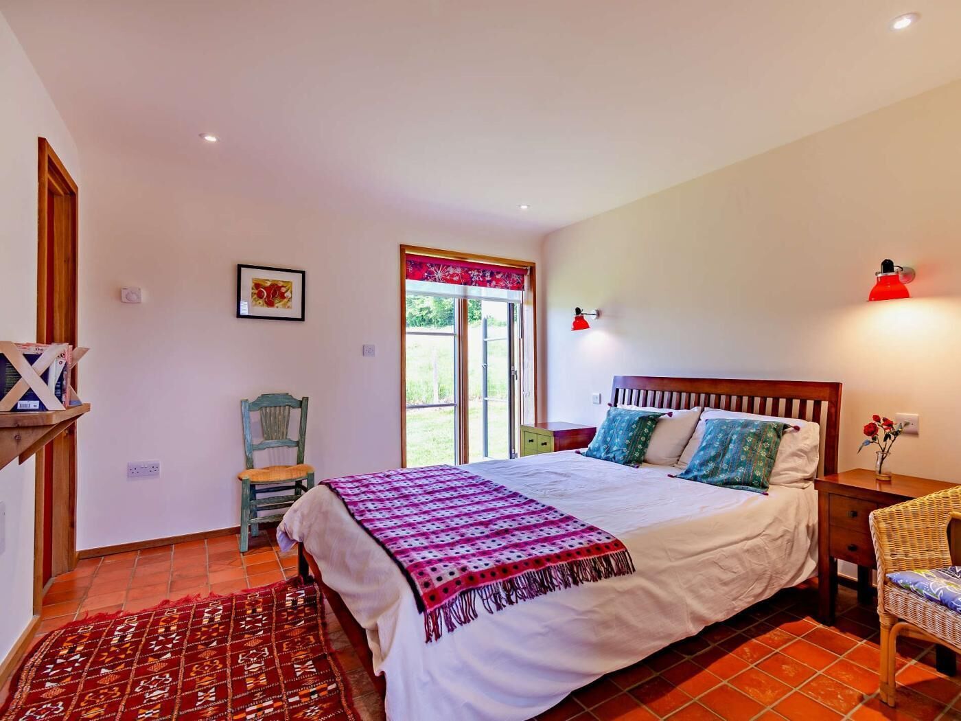 Un dormitorio con cama, una silla, una mesita de noche, una lámpara y una ventana.