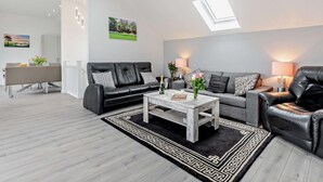Een moderne woonkamer met een zwarte leren bank, een witte salontafel en een grijze bank. Boven de grijze bank bevindt zich een dakraam, en aan de muur hangt een schilderij.