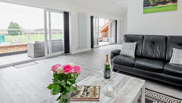 Een ruime woonkamer met een zwarte leren bank, een bijzettafeltje met een boek en bloemen, en uitzicht op een golfbaan door grote schuifdeuren.
