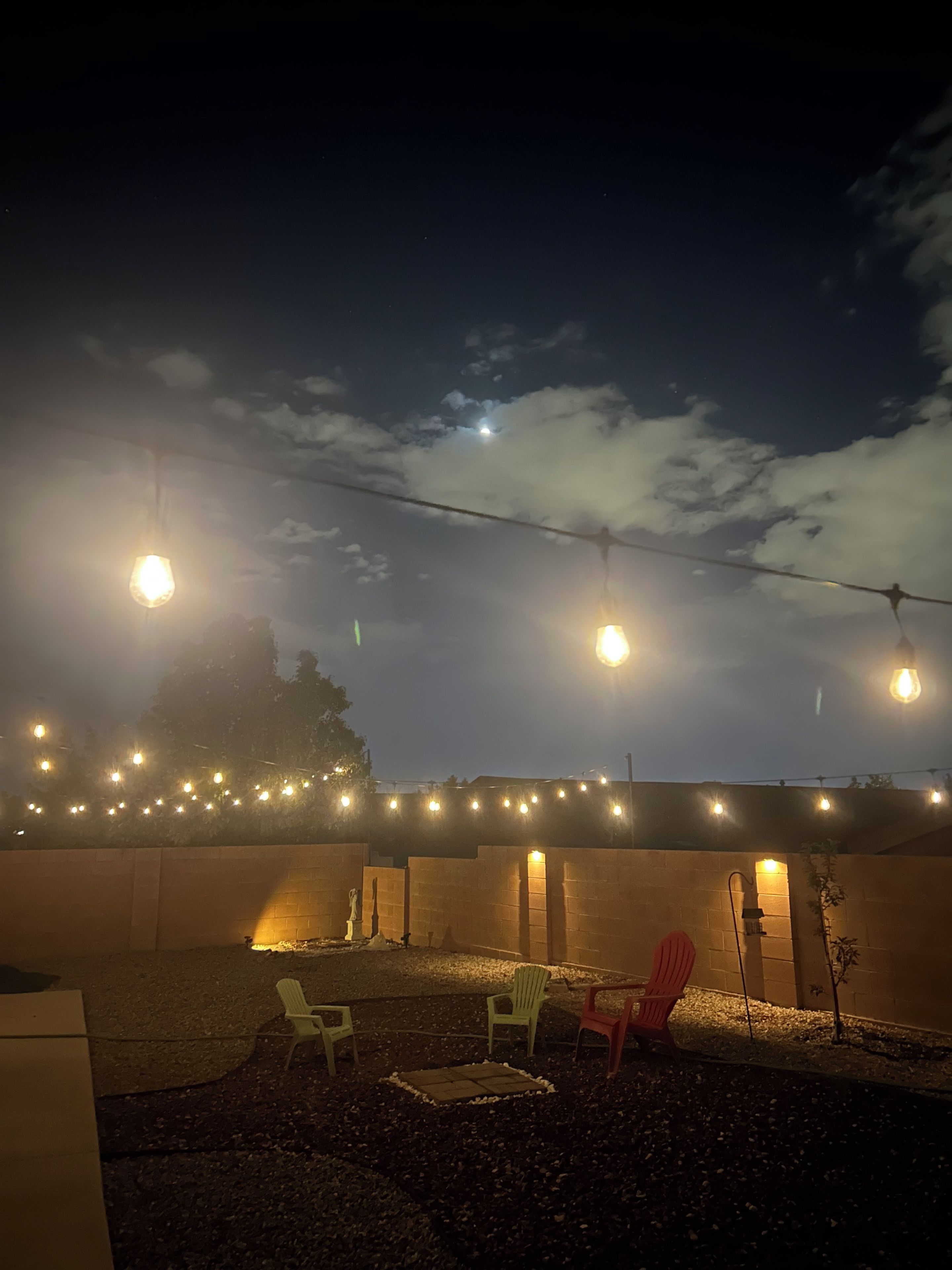 An outdoor seating area with chairs and a table, illuminated by string lights against a night sky with clouds.