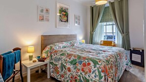 Une chambre à coucher comprenant un lit, une table de chevet, une chaise, une fenêtre avec des rideaux et des œuvres d’art murales.