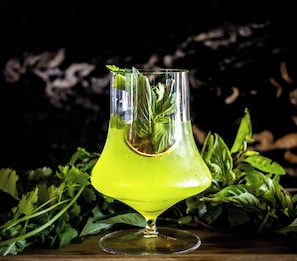 Un verre transparent contenant un liquide vert, peut-être un cocktail, et quelques feuilles de menthe à l’intérieur, posé sur une surface en bois, entouré d’herbes vertes.