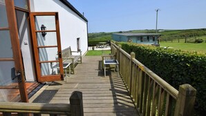 Eine Holzterrasse führt zu einem weißen Gebäude mit Glastür, einer Bank und einem Tisch.
