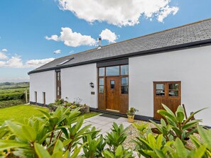 A white building with a grey roof, wooden doors, and windows, surrounded by greenery and a clear sky. - 4 Bed in Bideford (oc-owlbr) (Higher Clovelly)