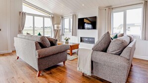 A spacious living room with two grey sofas, a wooden coffee table, a flat-screen TV, and large windows offering a view of the outdoors. - 2 Bed in St. Andrews (oc-93229) (St Andrews)