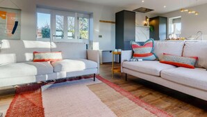 A modern living room with two sofas, a coffee table, and a rug. There is a kitchen area with a sink and a window. - 2 Bed in Arbroath (oc-84649) (Carmyllie)