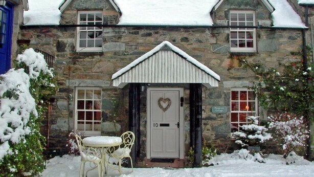 A stone building with a white door, two windows, and a small porch with a chair. - 2 Bed in Birnam, Dunkeld (oc-ca266) (Birnam)