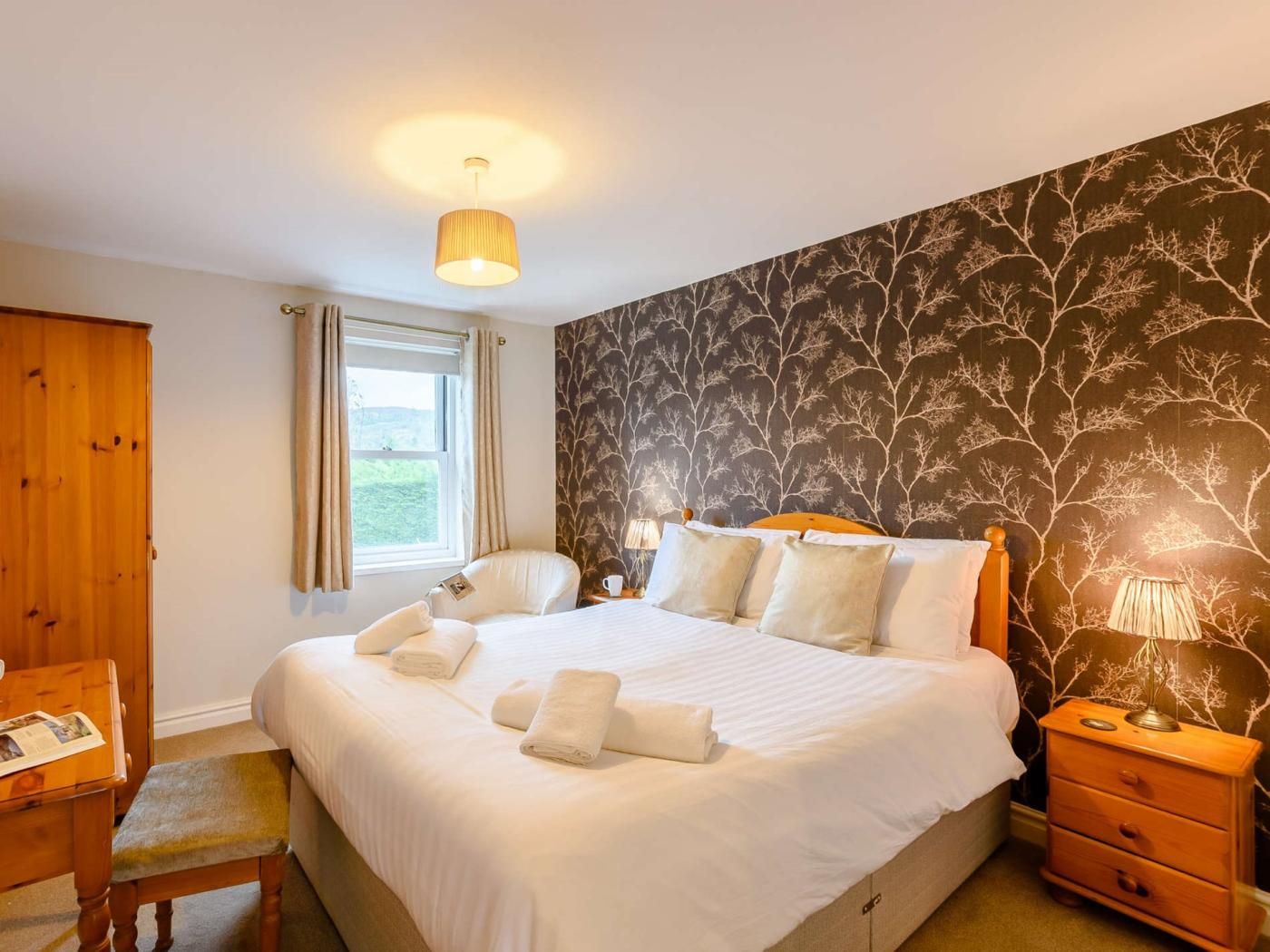A hotel room with a large bed, a wooden wardrobe, a window with curtains, a nightstand with a lamp, and a wall with a leaf pattern.