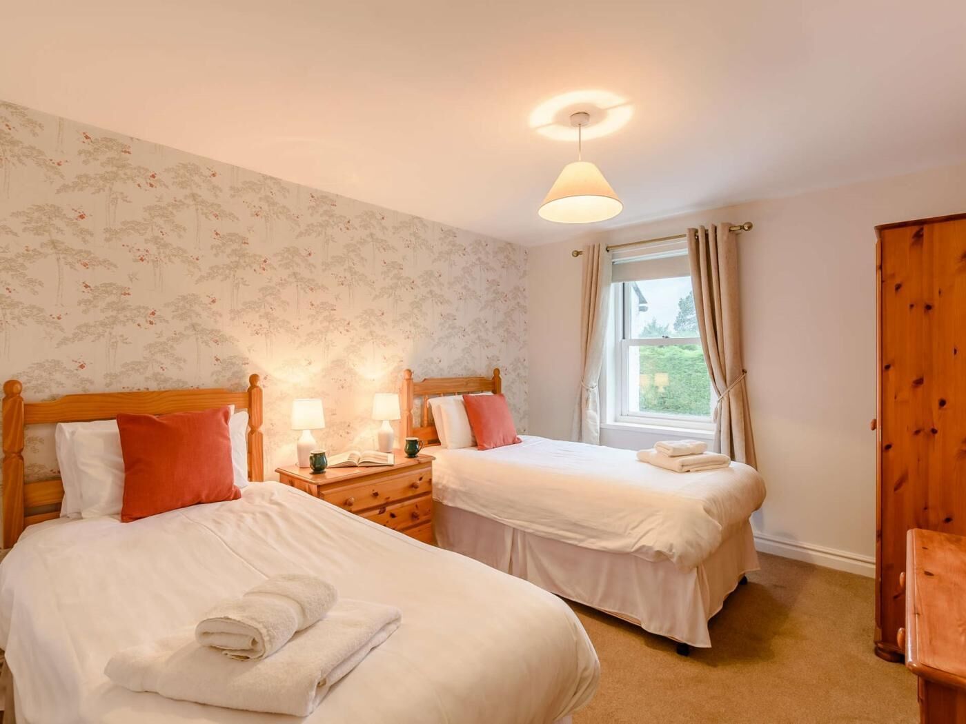 A bedroom with two beds, a nightstand with lamps, a window with curtains, and a wooden wardrobe.