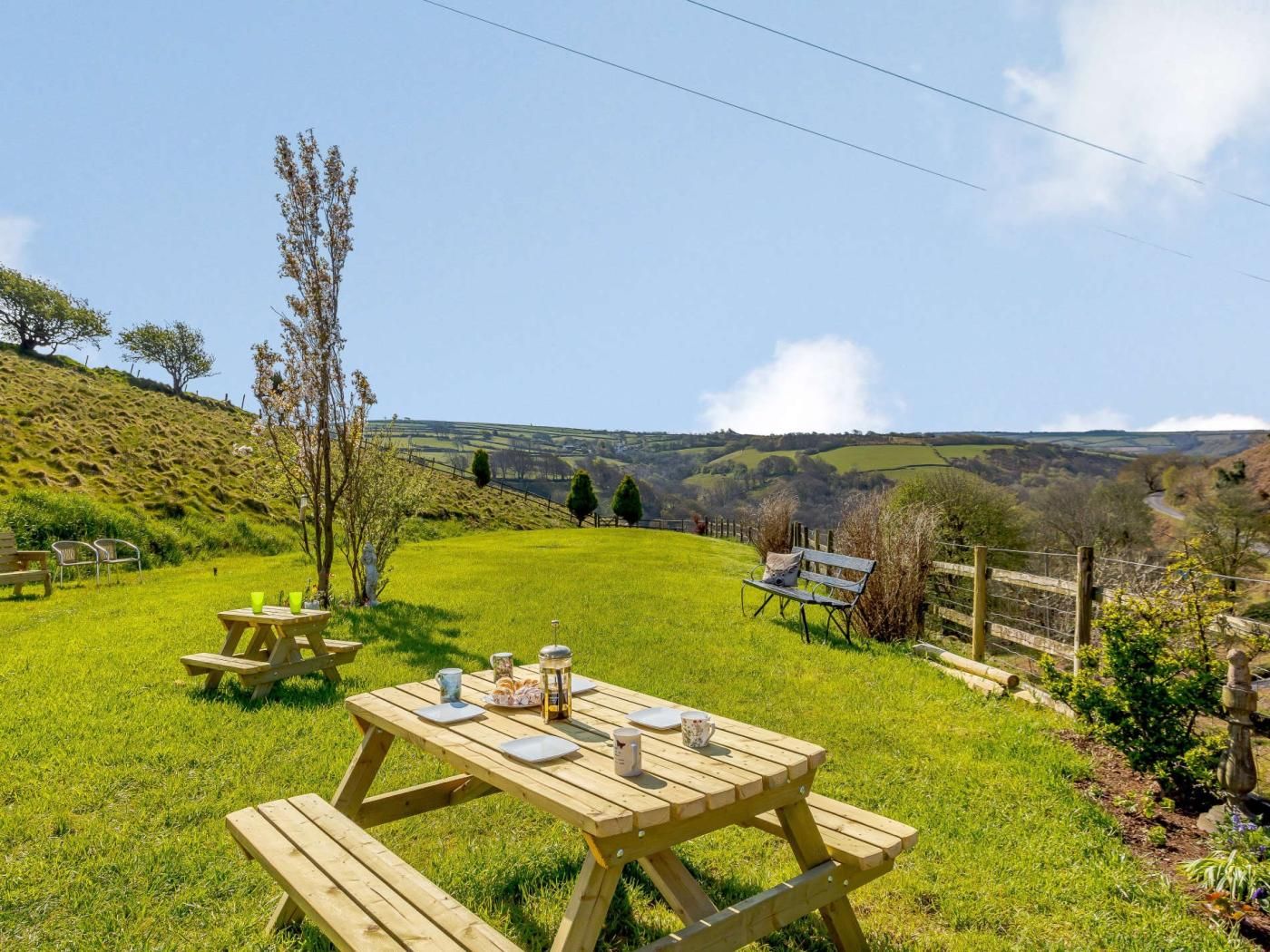 Un espace pique-nique avec des bancs et des tables en bois, entouré d’un champ verdoyant et offrant une vue sur des collines ondulantes.