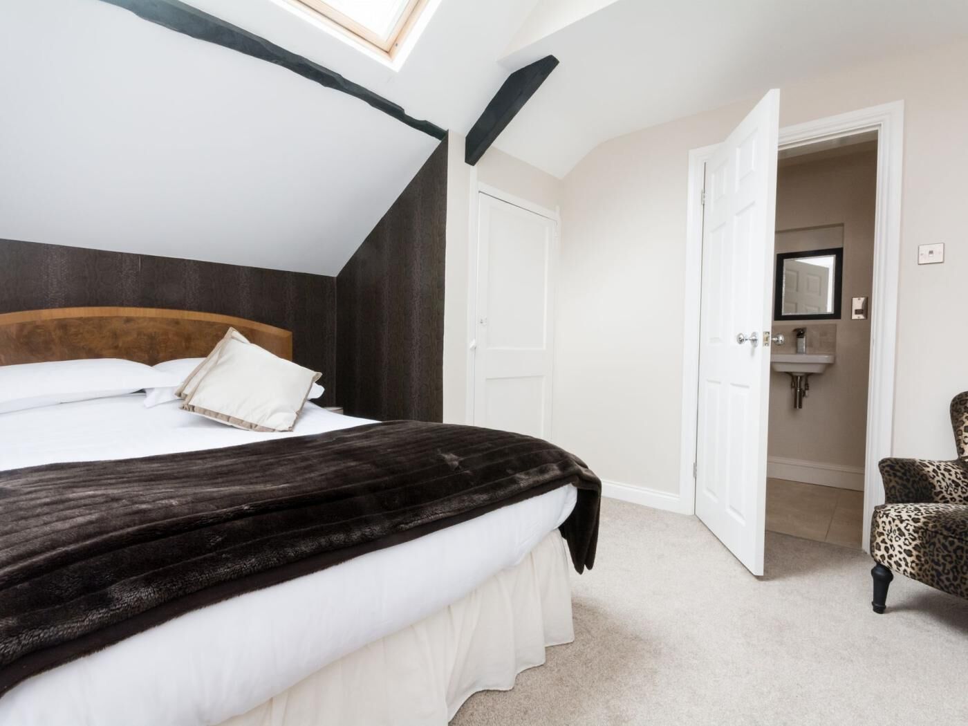Une chambre à coucher avec un lit, une lucarne, une porte donnant sur une salle de bain et un fauteuil à motifs léopard.
