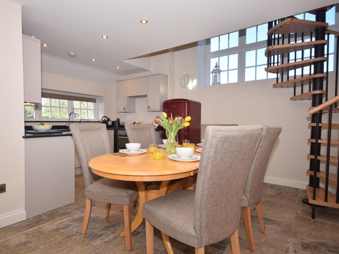 A dining area with a round table, four grey upholstered chairs, a vase with flowers, and a breakfast setting.