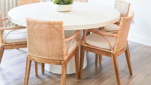 Une table de salle à manger ronde, entourée de chaises en osier, un lustre suspendu également en osier, et un tableau abstrait encadré accroché au mur.