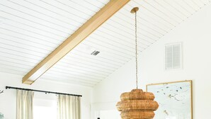 Un salon avec un canapé blanc, une table à manger, des chaises en osier, un luminaire suspendu et une œuvre d’art encadrée au mur.