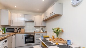 Een kleine keuken met een magnetron, oven en spoelbak, een eettafel met stoelen, en een klok aan de muur.