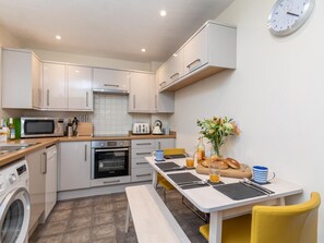A compact kitchen with a microwave, oven, and sink, a dining table with chairs, and a clock on the wall. - 2 Bed in Boscastle (oc-54474) (Boscastle)