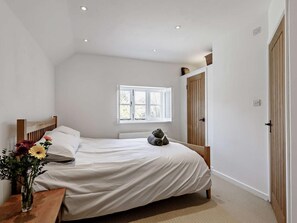 Un dormitorio con cama, ventana, puerta de madera y mesita de noche con un florero.