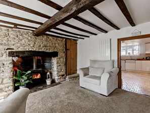 Ein gemütliches Wohnzimmer mit Kamin aus Stein, einem weißen Sessel und Blick in die Küche.