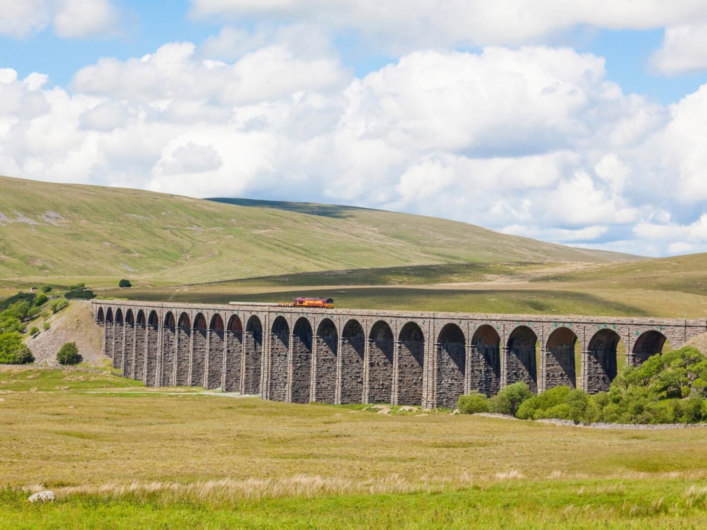 Un viaducte de pedra amb un tren creuant-lo, envoltat de camps i turons verds.