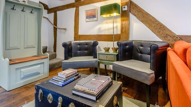 Ein gemütliches Wohnzimmer mit offenem Dachboden, einer grünen Kommode, zwei Ledersesseln, einem kleinen Tisch mit Büchern und einem Vintage-Koffer.