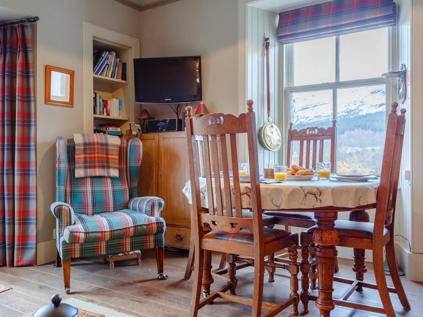 A cozy room with a plaid sofa, wooden chairs, a dining table with breakfast, a bookshelf, and a TV.