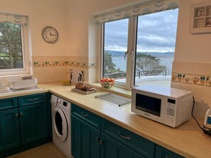 A kitchen with a washing machine, a microwave, and a window with a view of the sea. - 2 Bed in Aberdovey (oc-94565) (Aberdovey)