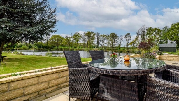 Un patio al aire libre con mobiliario de mimbre, una mesa de vidrio y vistas a un lago y árboles.