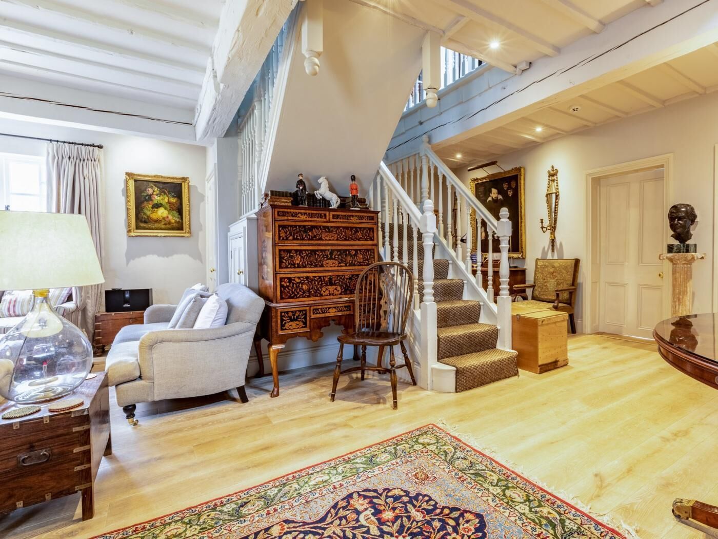 A spacious living room with a staircase, a wooden chest of drawers, a patterned rug, and a chandelier.
