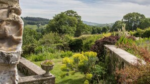 Edifici de pedra amb vista a un jardí, incloent diverses plantes i un paisatge allunyat.
