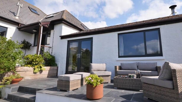 Une terrasse extérieure moderne, dotée de meubles en osier, d’une grande fenêtre et de plantes en pot.