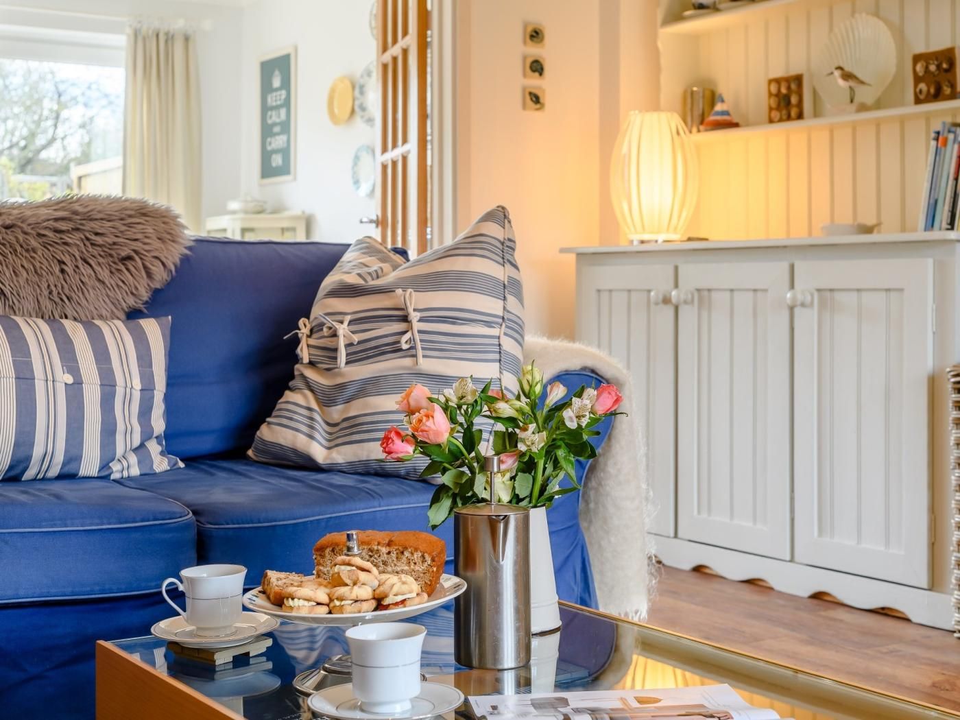 A cozy living room with a blue sofa, striped pillows, a vase with flowers, and a tray with pastries and cups.