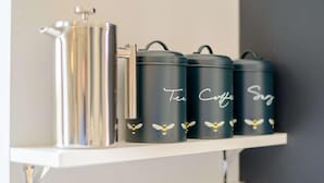En hylla med en silverfärgad behållare och tre svarta behållare märkta med orden ”Te”, ”Kaffe” och ”Sirap”.