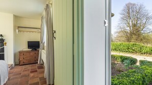 A bedroom with a bed, a television, a dresser, and a view of a garden through an open door. - 3 Bed in King's Lynn (oc-kt097) (Ingoldisthorpe)