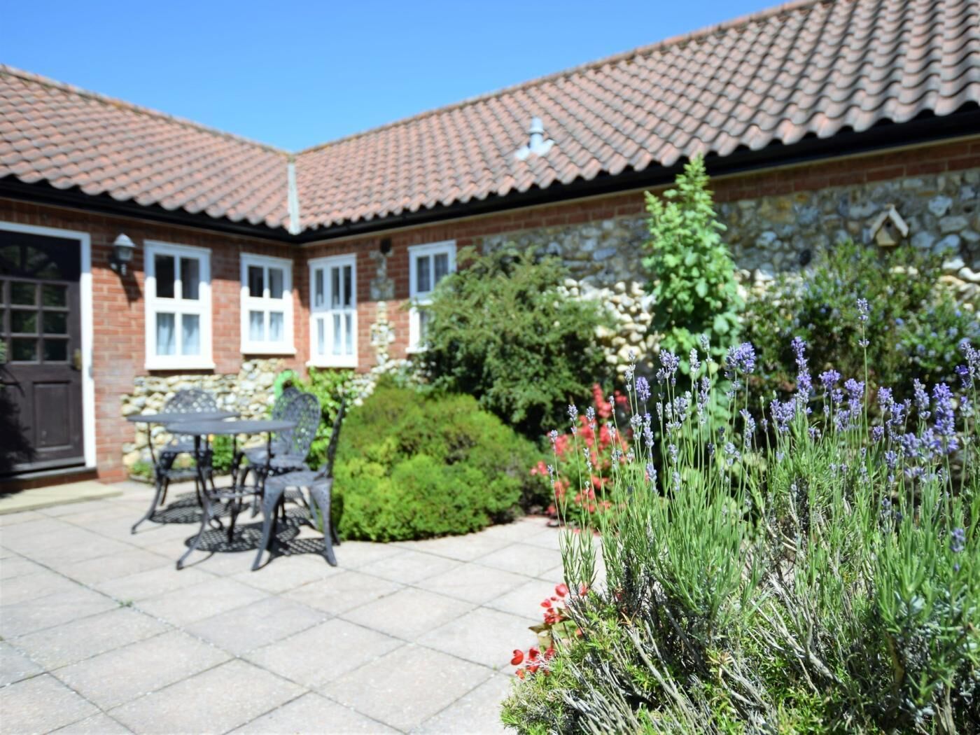 A brick house with a tiled roof, a patio with a table and chairs, and a garden with lavender and red flowers.